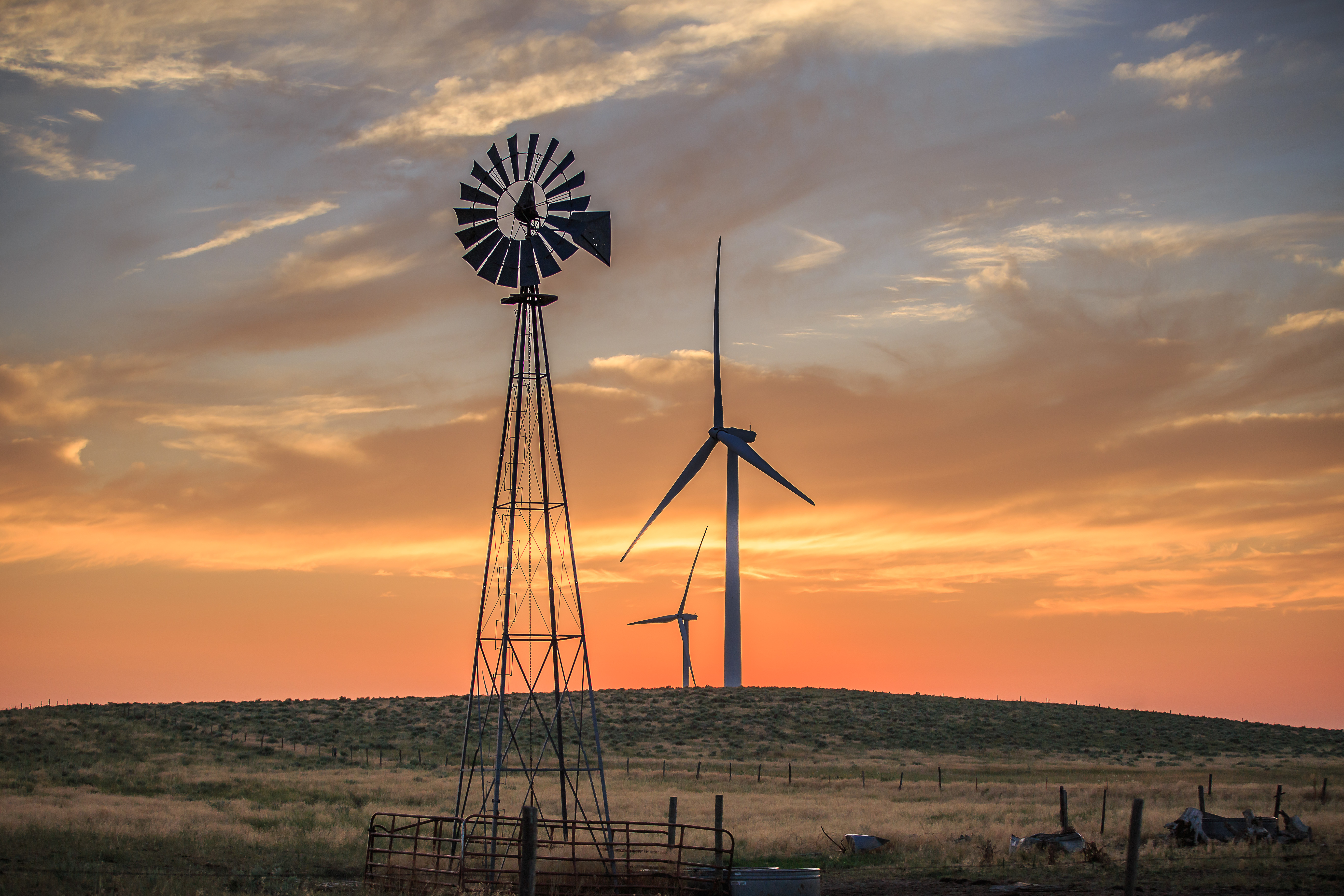 A weather vane and two wind turbines are positioned on a hill with the setting sun behind them.