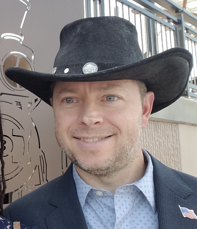 Sam Anderson stands in front of a balcony with a black cowboy hat and a suit. 