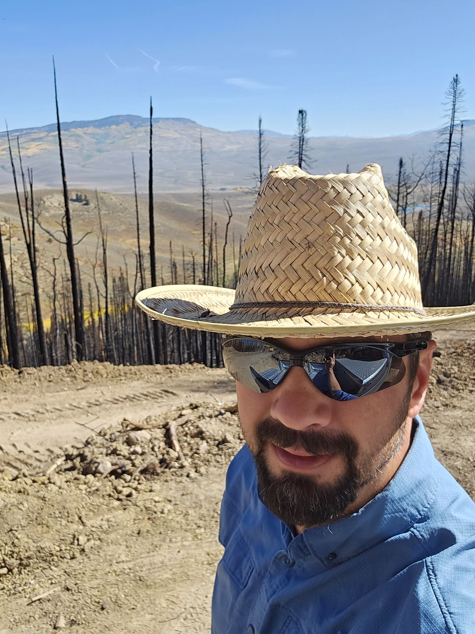 John is standing on a dirt road with burnt trees and mountains in the background