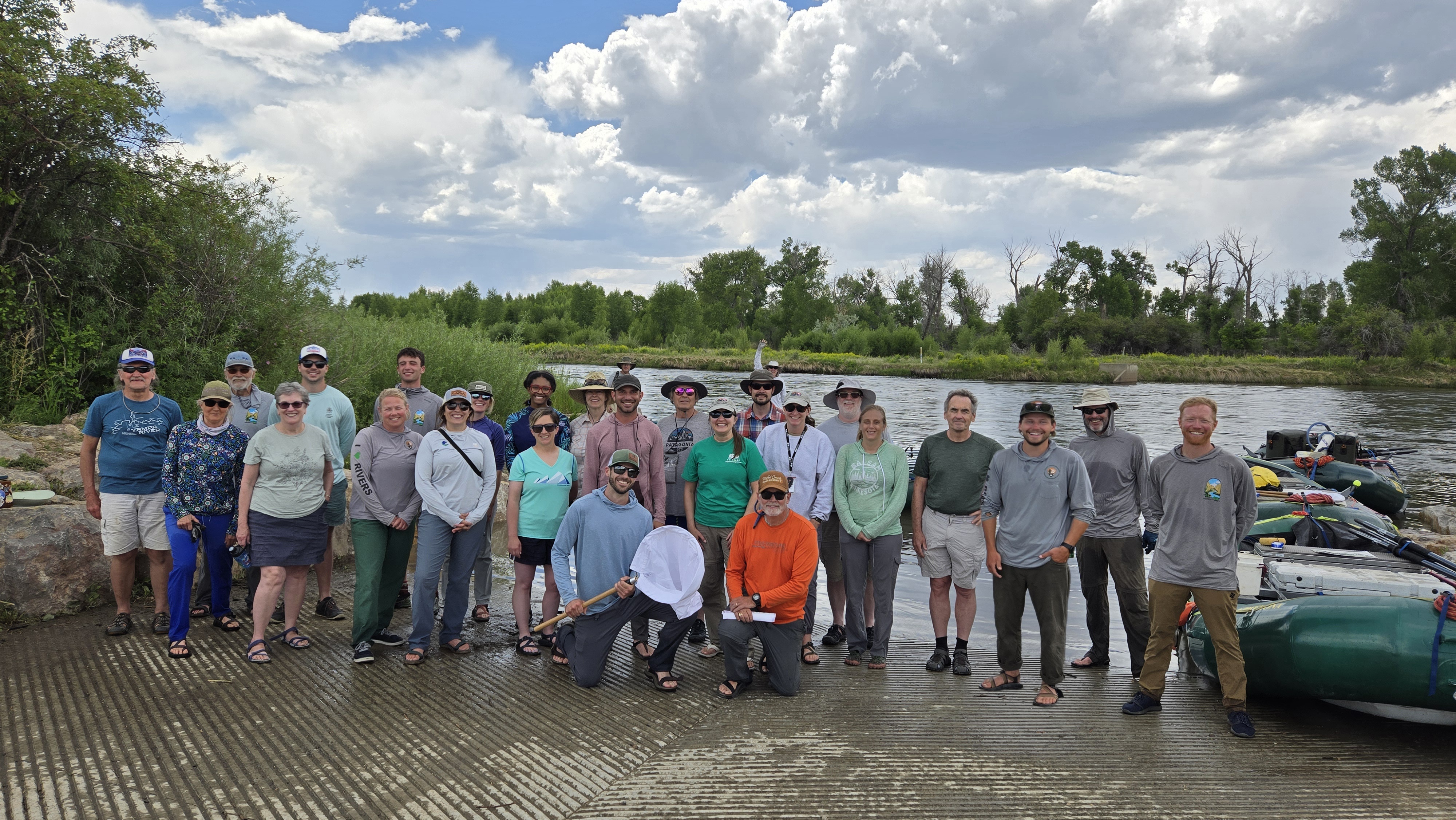 A group of 25 people poses for a photo on a boat landing with multiple rafts on the right side, and the Yampa River in the background