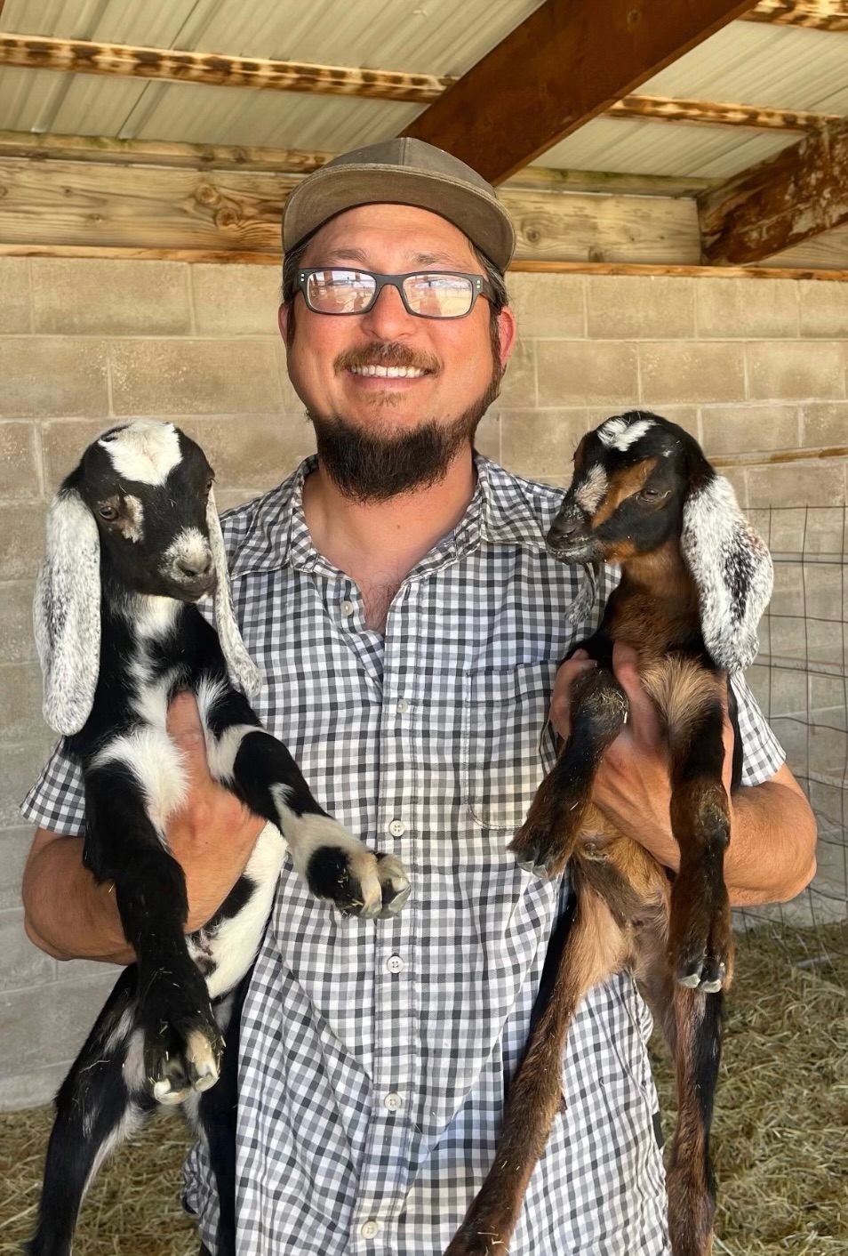 Anthony wears a baseball hat and glasses while holding two small goats