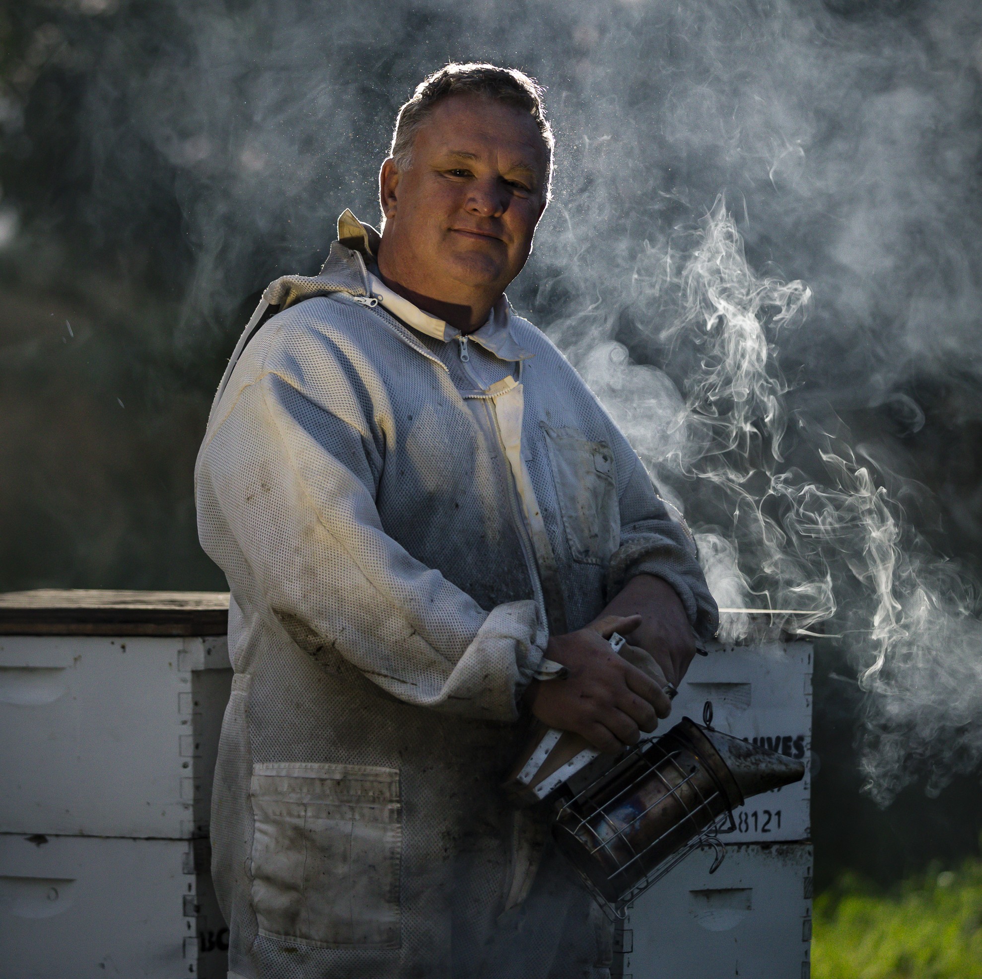 Portrait of Robert Bishop-Cotner standing in his beekeeper suit in the smoke