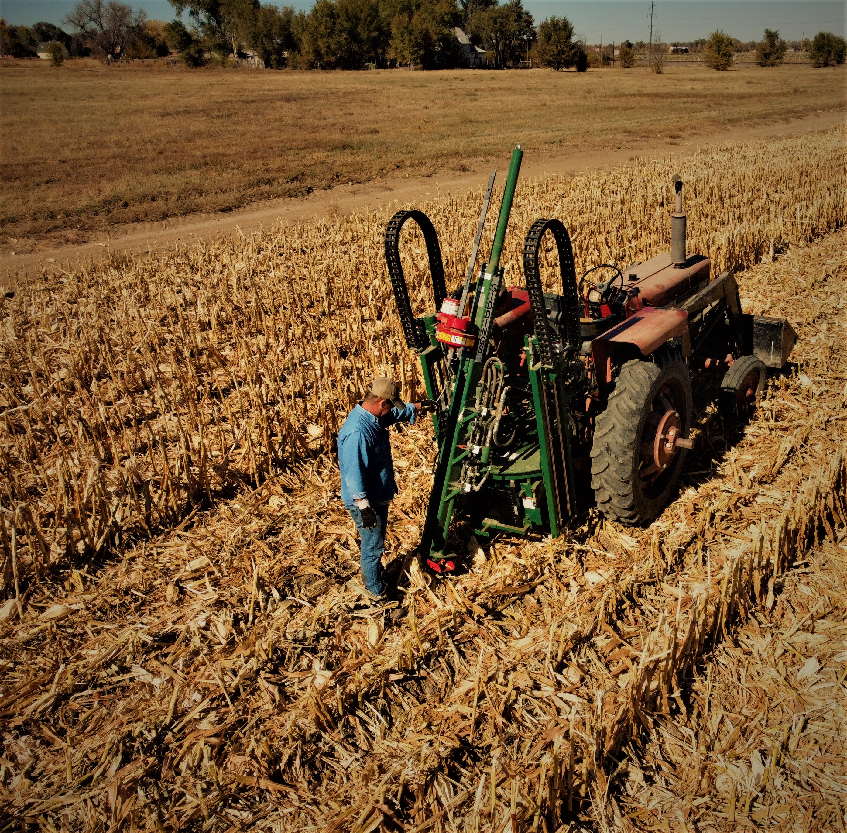 A tractor and implement are used to take a soil sample in a corn field as a farmer looks on
