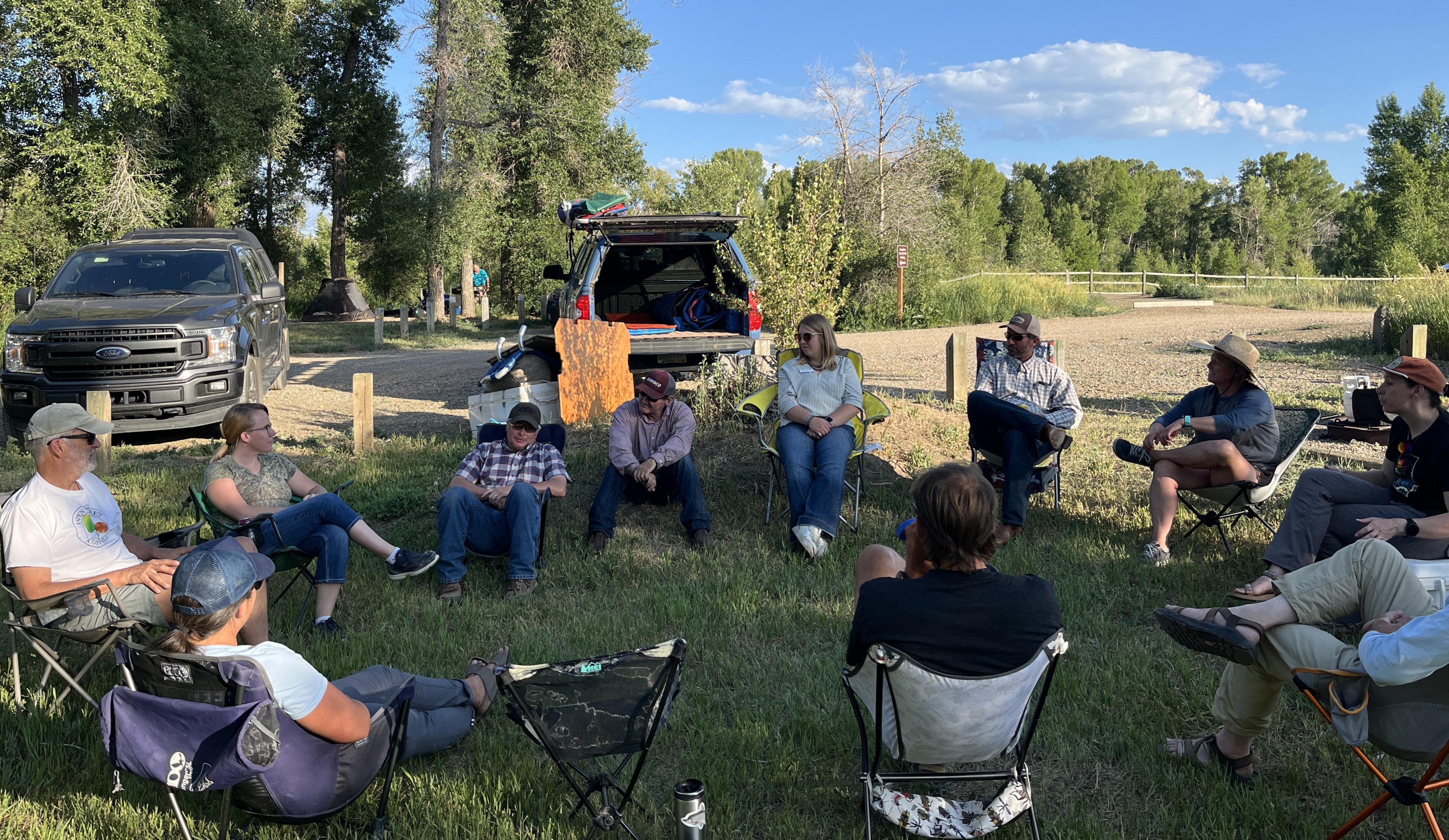 A group sits in camping chairs in a circle in a shady state park