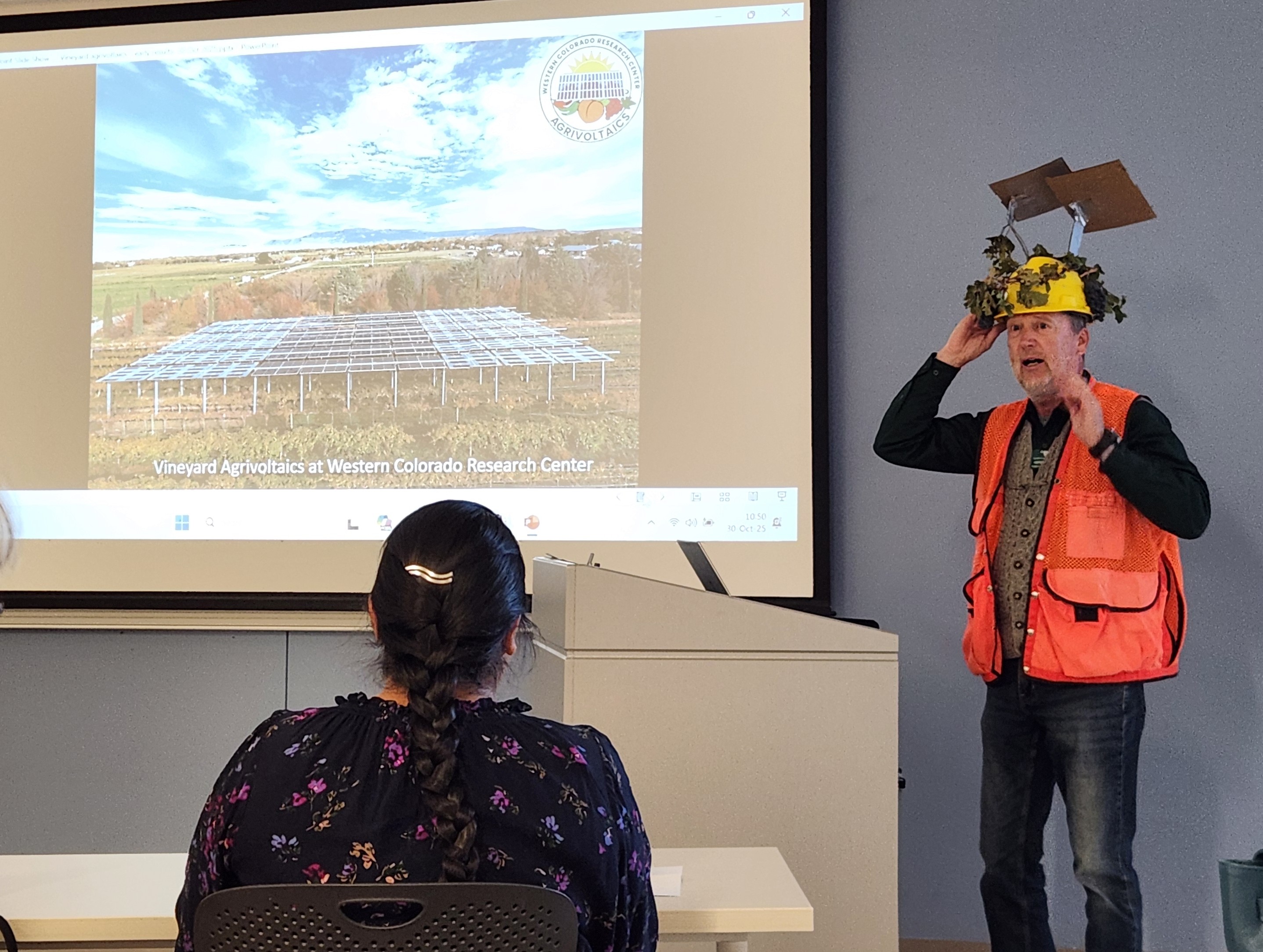 Man in a hard hat with solar panels and grapes on it presents in front of a screen