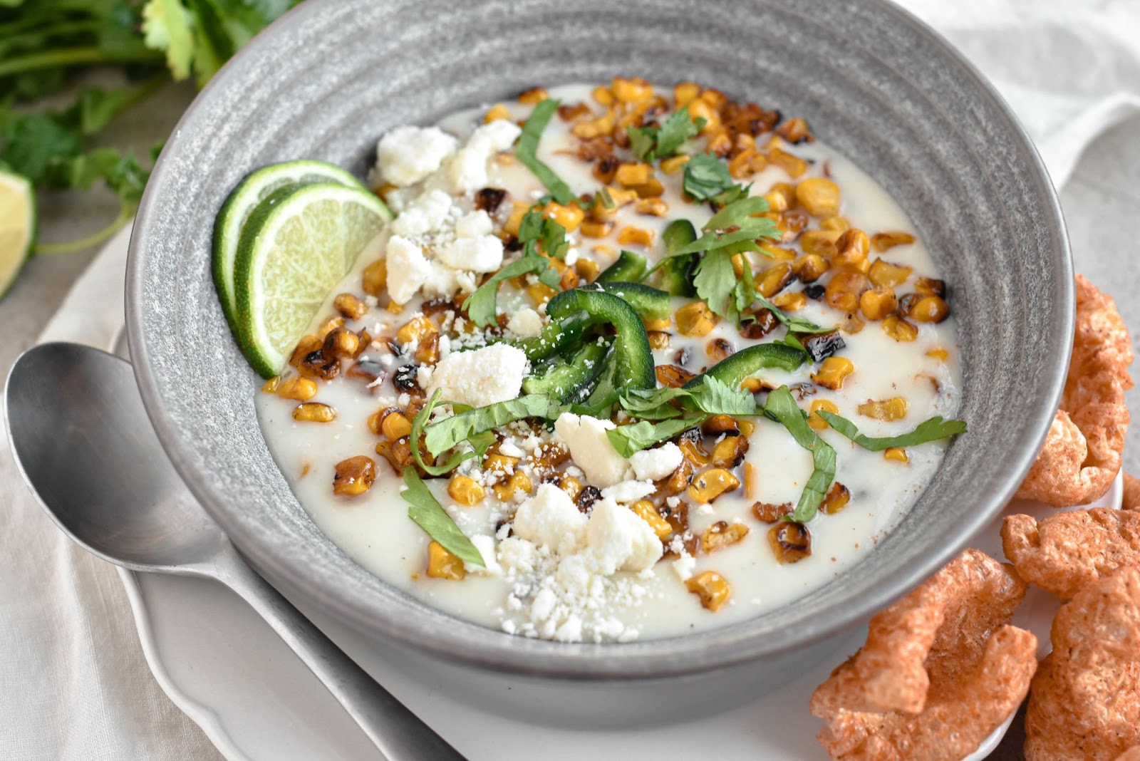 A bowl of creamy queso topped with roasted corn, sliced limes, cilantro, cheese crumbles, and jalapeño slices, accompanied by crispy pork rinds on the side