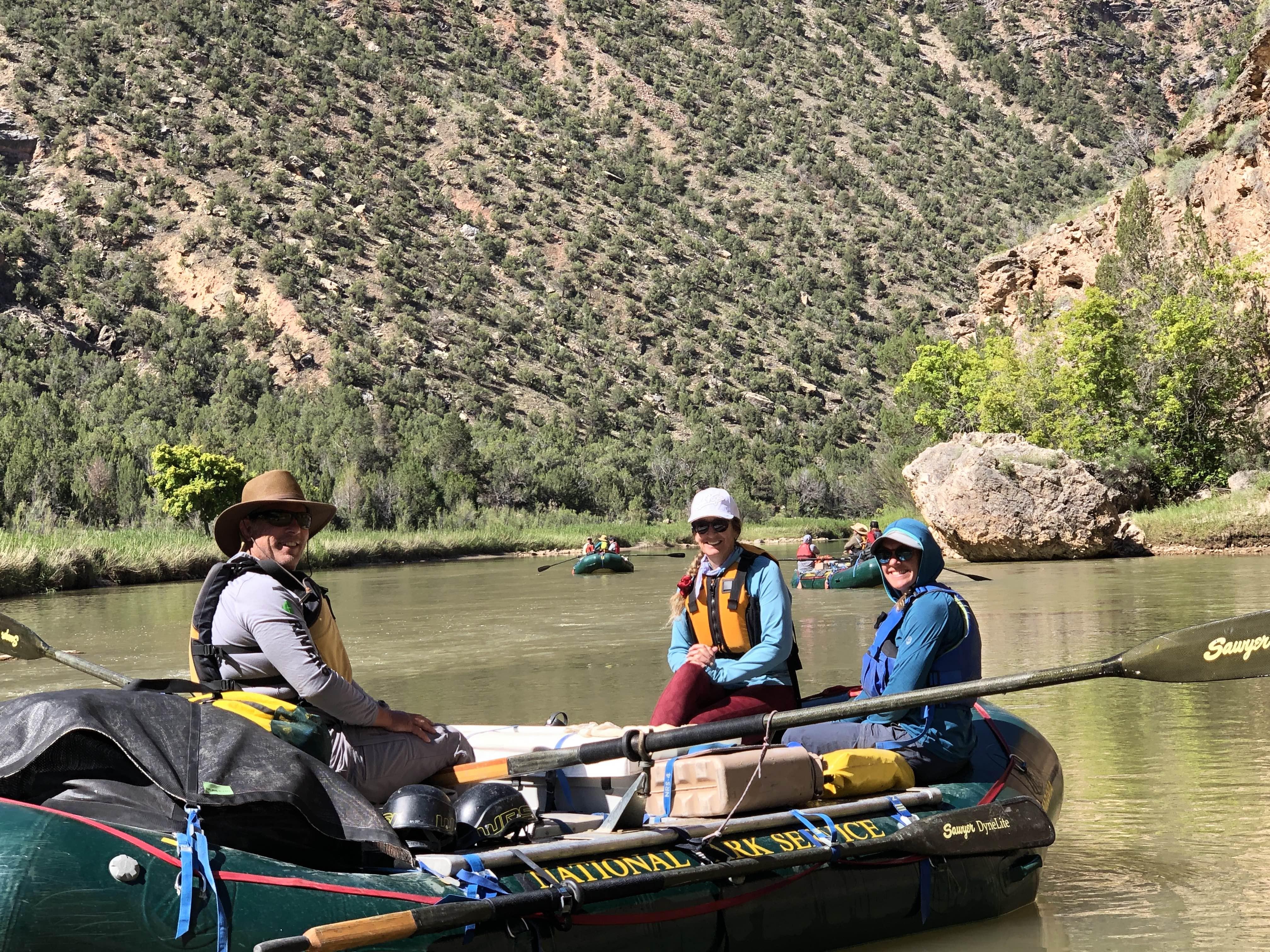 Two women and one man sit in a raft posing for a picture on the Green River, with other rafts on the river in the background. 