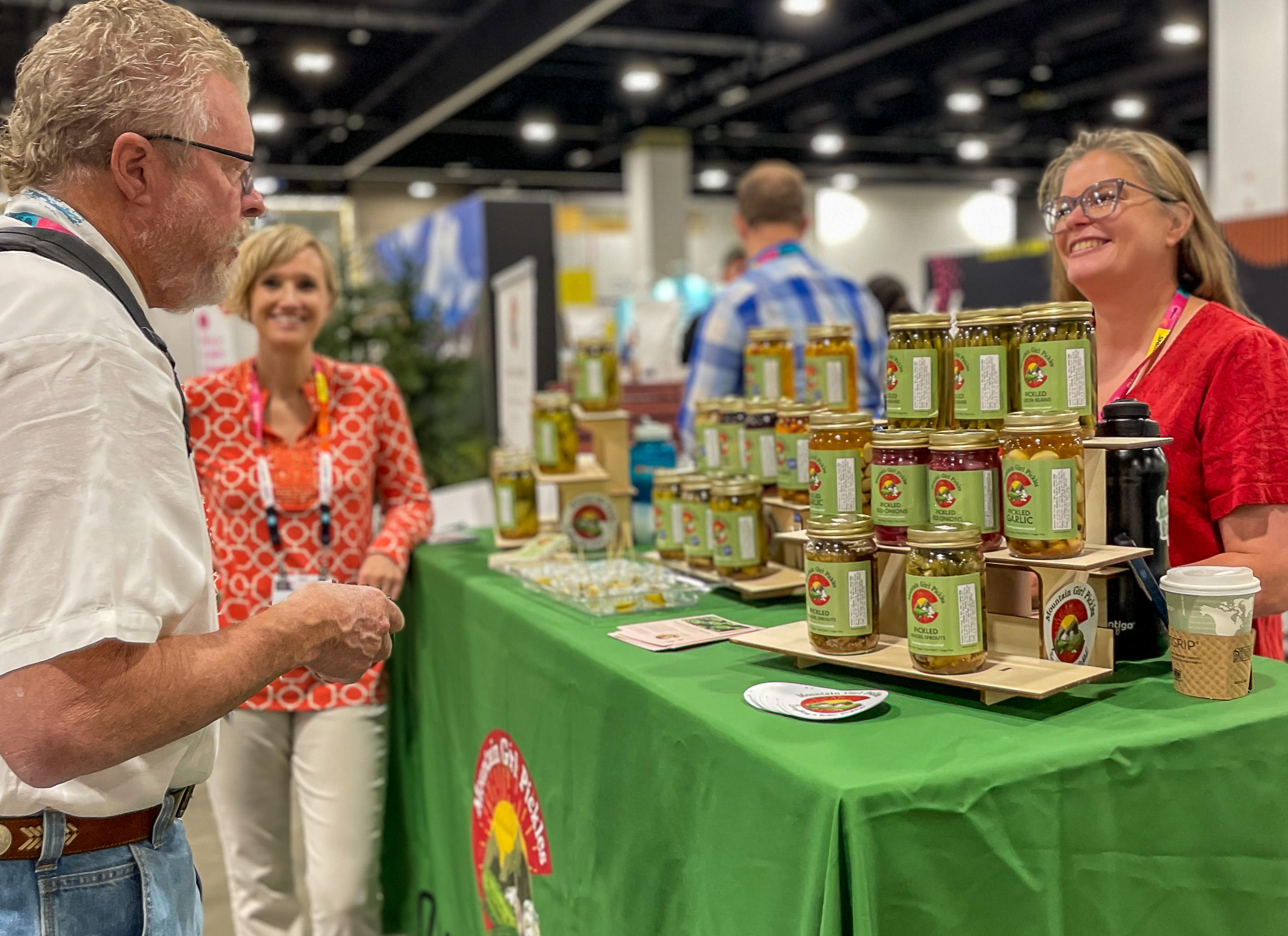 People gather around stacked jars of Colorado-made salsa on a green tablecloth at a trade show