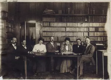 Five men and two women sit facing the camera at a table in front of book shelves circa 1907.