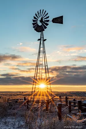 The sunset's rays burst through clouds and the legs of a windmill standing tall against the dim lit blue sky, photo by Tim Atkinson.
