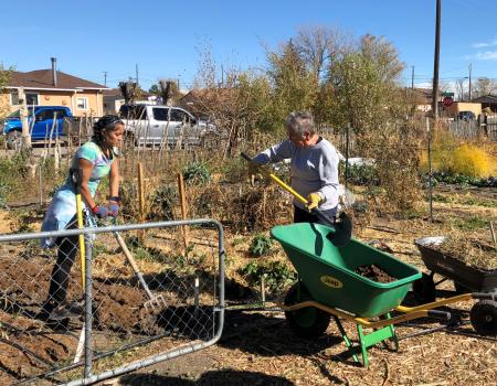 A woman with a four prong pitchfork is tilling soil while another person shovels dirt into a wheelbarrow at the Earth Mountain Farm Community Garden