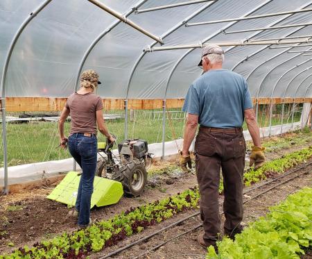 Brightwood Farms: a female farmer in a greenhouse using machinery is being watched by an older farmer.