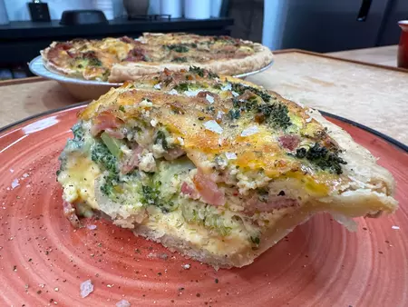 Colorado Proud Quiche: a slice of quiche with broccoli, bacon, cheese and flaky salt on a red plate with the remainder of the quiche in the background